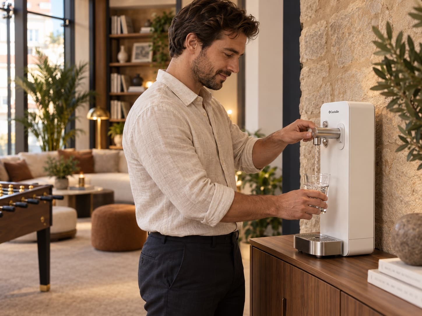 La fontaine à eau Castalie de Maison Voyna, posée sur un buffet de noyer contre un mur de pierre : un membre remplit un verre d'eau pétillante, le salon et le babyfoot derrière lui.