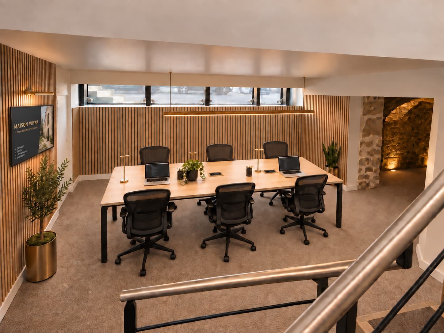 The long table at Maison Voyna, at the foot of the vault: six ergonomic chairs around a pale wooden surface, oak panelling, a linear pendant light, a band of windows at pavement level and the lit stone arch at the back.