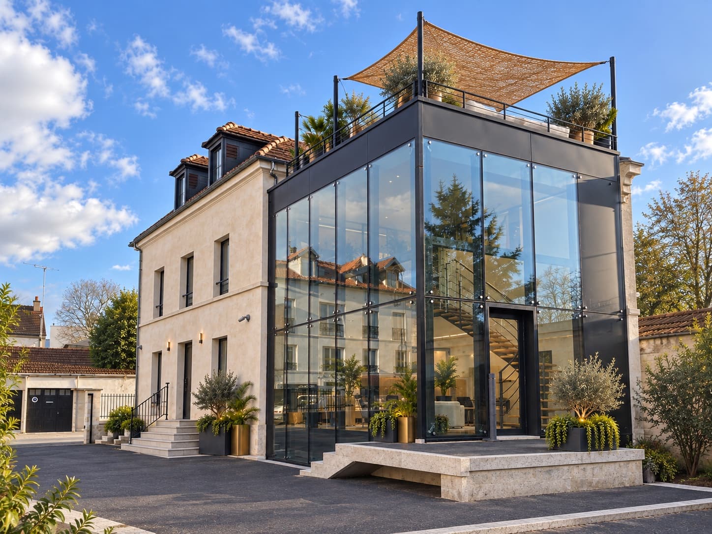 Maison Voyna vue de la rue : la maison de maître en pierre claire et, greffée contre son flanc, l'extension vitrée à ossature d'acier noir surmontée de la terrasse et de sa toile d'ombrage.